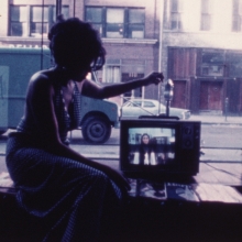 Old photograph of a person sitting next to a small tv. In the background are building and cars parked on the street.