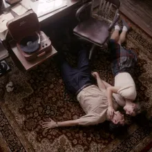 Two people lying on the floor in an office with dark brown hard wood floors, dark brown wooden chairs. One person is wearing long dark pants with a tan short sleeve button up shirt. The other person is wearing a dark plaid skirt with a white shirt sleeve top, ankles socks, with dark brown shoes.