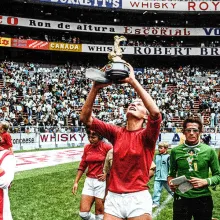 Film still from 'Copa 71'. Woman soccer player holding up World Cup trophy in a stadium filled with fans and her team on the field with her.