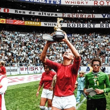 Film still from 'Copa 71'. Woman soccer player holding up World Cup trophy in a stadium filled with fans and her team on the field with her.