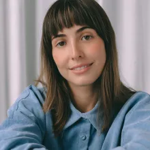 A woman with white skin smiles at the camera. She has straight brown hair, bangs, and brown eyes and is wearing a light blue button-down shirt.