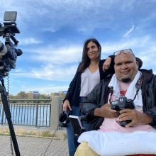 Jay, a Dominican man with a goatee and reading glasses perched on top of his head, sits in a wheelchair holding a digital camera. Alexis, a white woman with dark brown hair stands next to him but a bit behind, holding headphones attached to a large camera on a tripod. They are smiling softly, looking at the camera, and wearing light jackets on a boardwalk in front of a river on a sunny day with clouds in the blue sky.