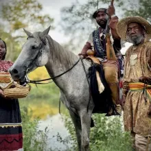 Three Black Seminoles, a woman and two men, wearing traditional clothing. One of the men is sitting on a gray horse while the others stand next to the horse.