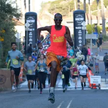 A man wearing sunglasses, shorts, and a neon colored top sprints ahead of a group of runners on a paved street