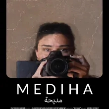 film poster for 'Mediha.' Black and brown background with white text. Image of a person holding up a video camera. 