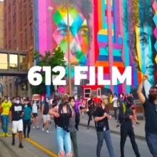 Thousands of protestors dressed in summer clothes march in front of a colorful mural 