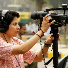 A Brown, badass, butch dyke is shooting a scene with queer rights protesters (offscreen) on the streets of New Delhi, with a video camera on a monopod and headphones covering her ears.