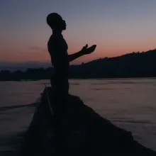 Film still from 'Eat Bitter,' showing the silhouette of a man with arms outstretched, on a small boat.