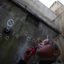 A young girl with hair in a ponytail blows bubbles in front of a large, dark building