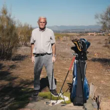 Portrait of an older man in a field, he has a golf club in his hand and next to him is the rest of the kit. He looks at the camera concentrated. The landscape is like a desert.