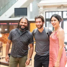 Three people standing in a brightly lit room, attending Getting Real '22. (left to right) Man is wearing a black and gray shirt and olive green pants, man in middle is wearing a backpack with a gray shirt and black pants, and woman on the right is wearing a pink tank top dress with her brown hair clipped in the back.