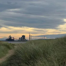 Landscape photo with tall green grass in the foreground with a sand path that leads to a river. Off in the distant background is a processing factory. The cloud filled sky with bits of sun poking through fills the rest of the landscape.