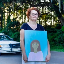 White woman with short red hair and glasses holds in front of her body a painted self-portrait of herself as a young girl while a police car drives behind her on a rural highway. Sun is glimmering in through trees in the background. In the painting she is wearing a pink shirt and is in front of a light blue background.