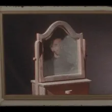 Ricardo, a pale man with glasses, dark short hair and a beard, is in the middle of the frame reflected and obscured in the mirror of a vintage vanity. The scene is framed by a window and the whole image has the soft quality of film.