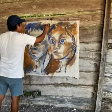 Artist Horazio Sanchez wearing a white shirt and shorts, with a blue hat on his head stands in front of a painting of two teenagers from the Calakmul Biosphere Reserve holding a paintbrush in one hand and applying it to the canvas.