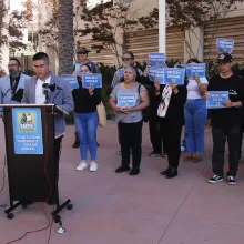 A group of protesters gathers behind a podium holding signs that say "Democracy for All" in English,Spanish and Vietnamese.