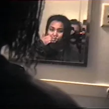 VHS still frame looking over the shoulder of someone in her early 20s with shoulder length dark hair into the mirror reflection where her hand is covering her mouth. A second person in her early 20s is visible in the mirror with a camcorder in front of her face.