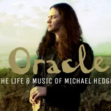 A middle aged man with long hair and a guitar stands on the coastal bluffs of California.