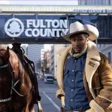 A man wearing a wide brim hat and a shearling jacket stands next to a brown horse in front of the Fulton County Courthouse
