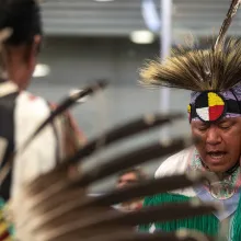 A man on the right is wearing an elaborate white, red, yellow, black, and purple beaded headdress while drumming at a ceremony at the Trout Lake Pow Wow. A blurry image of another man dressed in traditional attire with his back turned to his left.