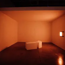 Total Running Time includes multiple works situated clockwise in a warmly lit alcove. Rescue, a plaster wall relief, and two geometric forms are positioned on the gallery floor. A niche on the rightmost wall contains two manipulated light sources, one of which creates a daylight effect.