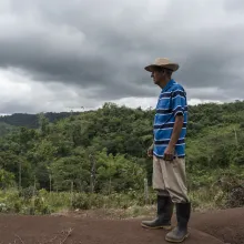 A man wearing a hat and rain boots stands in a hill, in front of some woods. There are dark clouds above