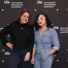 Two IDA members standing in front of a black step and repeat with white ida logos. Person on left is wearing all black, person on right is wearing a blue jean jumpsuit with glasses.