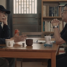 Two figures wearing dark shirts and glasses sit at a square table. They are surrounded by bookshelves, and are each drinking a mug of tea.