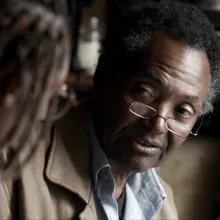 An older Black man wearing glasses is interviewing a blurry figure in the foreground