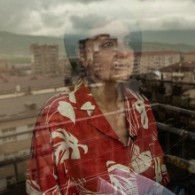 A woman wearing a patterned red shirt stands in a window, which shows a reflection of a rugged mountainous landscape.