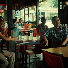 A couple sit in a dark restaurant, looking up at something out of frame.