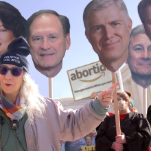 Still image from 'No One Asked You,' depicting a white woman with long blond hair in front of a protest holding large cardboard cutout faces of US Supreme Court justices.