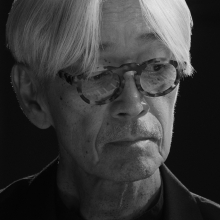 Still image from 'Ryuichi Sakamoto | Opus' depicting Sakamoto, an elderly Asian man wearing tortoiseshell glasses and with a fall of white hair over his forehead.