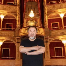 An Asian man wearing a black shirt stands with his arms crossed in front of the balcony seating of an opera house