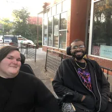 Kennedy, a white, fat, femme, and Justin, a Black man smile at the camera at an outdoor event. They both are dressed in black fall clothing and are seated in their power wheelchairs.