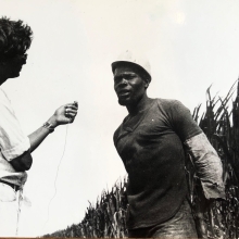 black and white photo from the Victor Jara Collective featuring a man holding a small microphone up to another man wearing a hard hat standing against a row of brush
