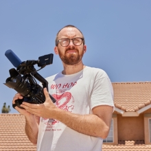 In a very bright, sunny driveway, a white man holds a video camera while squinting into the sun.