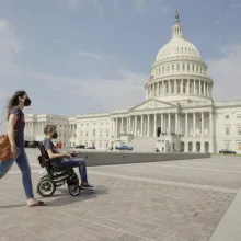 Two people outside a government building wearing masks. One person is in a wheelchair.