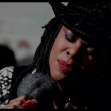 Still from Q Lazzarus performing in the movie “Philadelphia”. Q, an African-American woman, is wearing a black hat and holding a microphone she is singing into.