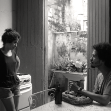Black and white still of a young couple looking at each other, in their kitchen.