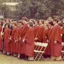 A large group of young men in red robes stand on the football field of their school, preparing for their graduation.