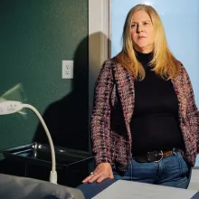 A middle-aged woman with long blonde hair, wearing a jacket and jeans, stands behind a medical exam chair.