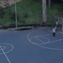 An African American man wearing a gray hoodie stands on an empty basketball court, reaching up to touch the basketball net