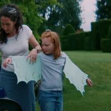 A woman helps a child put on paper wings. They are standing in a grassy field.