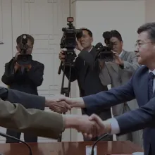 Two officials shake hands as a camera crew films them in the background.