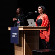 Two women stand onstage, one at the podium giving a keynote address.