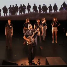 A man stands strumming a banjo in the middle of a stage, singing into a microphone. Other musicians and players surround him while “The Edge of Nature” plays on the screen behind them all.