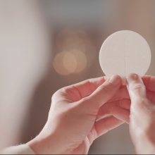 An over-the-shoulder of a pair of hands holding a small, white disc, the sacramental bread of the Eucharist.
