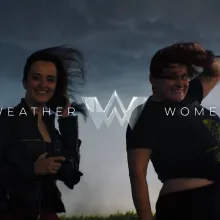 Two women standing outdoors against a backdrop of dark, stormy clouds, smiling. Their hair is being blown by the wind. The text "WEATHER WOMEN" is superimposed on top