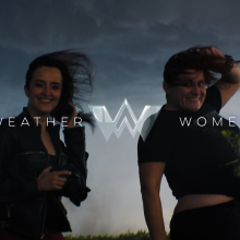 Two women standing outdoors against a backdrop of dark, stormy clouds, smiling. Their hair is being blown by the wind. The text "WEATHER WOMEN" is superimposed on top